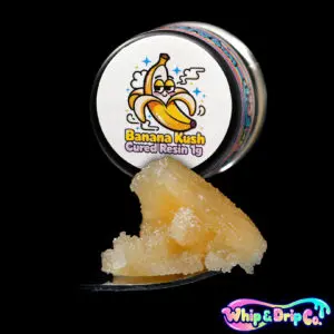 Whip & Drip Co. Banana Kush cured resin concentrate with pale golden extract displayed beside tropical banana-themed 1 gram product packaging on a black background.