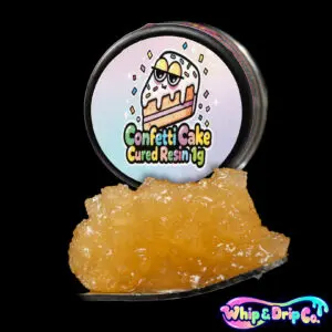 “Whip & Drip Co. Confetti Cake cured resin concentrate with golden extract displayed beside cake-themed 1 gram product packaging on a black background.”