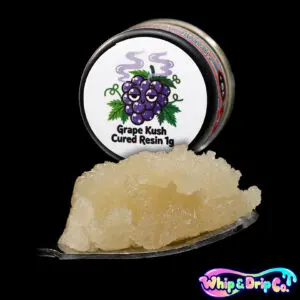 “Whip & Drip Co. Grape Kush cured resin concentrate with golden extract displayed beside grape-themed 1 gram product packaging on a black background.”