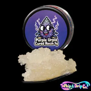 “Whip & Drip Co. Purple Urple cured resin concentrate with pale golden extract displayed beside purple grape-themed 1 gram product packaging on a black background.”