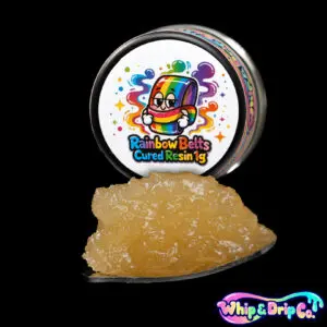 “Whip & Drip Co. Rainbow Belts cured resin concentrate with pale golden extract displayed beside colourful rainbow-themed 1 gram product packaging on a black background.”