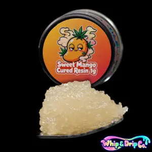 “Whip & Drip Co. Sweet Mango cured resin concentrate with pale golden extract displayed beside tropical mango-themed 1 gram product packaging on a black background.”