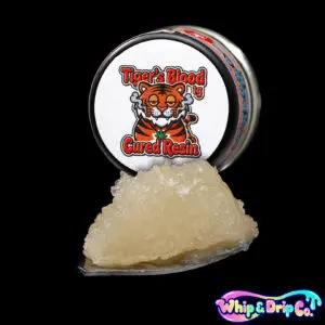 “Whip & Drip Co. Tigers Blood cured resin concentrate with golden extract displayed beside colourful tropical-themed 1 gram product packaging on a black background.”